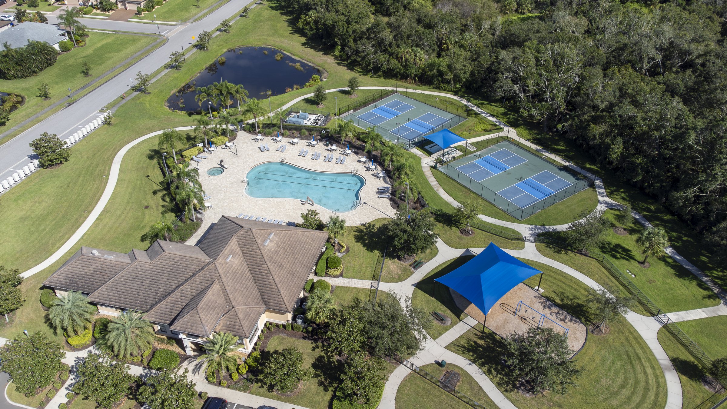 Wide community amenity park with pool, tennis, and shaded playground — Sarasota drone aerial